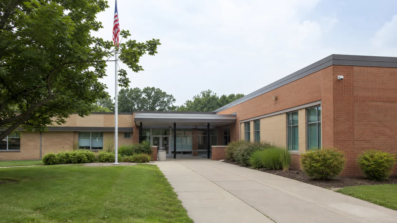 Public school campus entrance with brick building, flagpole, walkway, and landscaping.