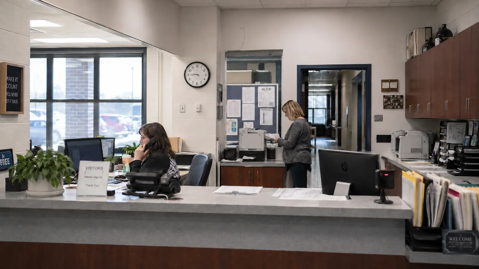 School front office staff working at a reception counter with office phones and administrative paperwork.